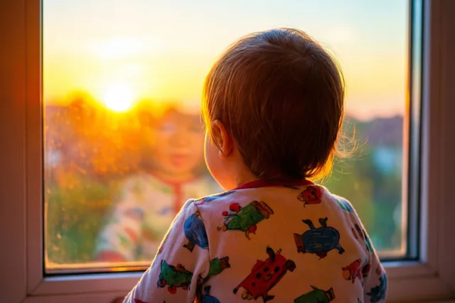 Een peuter in een pyjama met een patroon kijkt, van achteren gezien, door een raam naar een zonsopgang in warme gouden en oranje tinten.