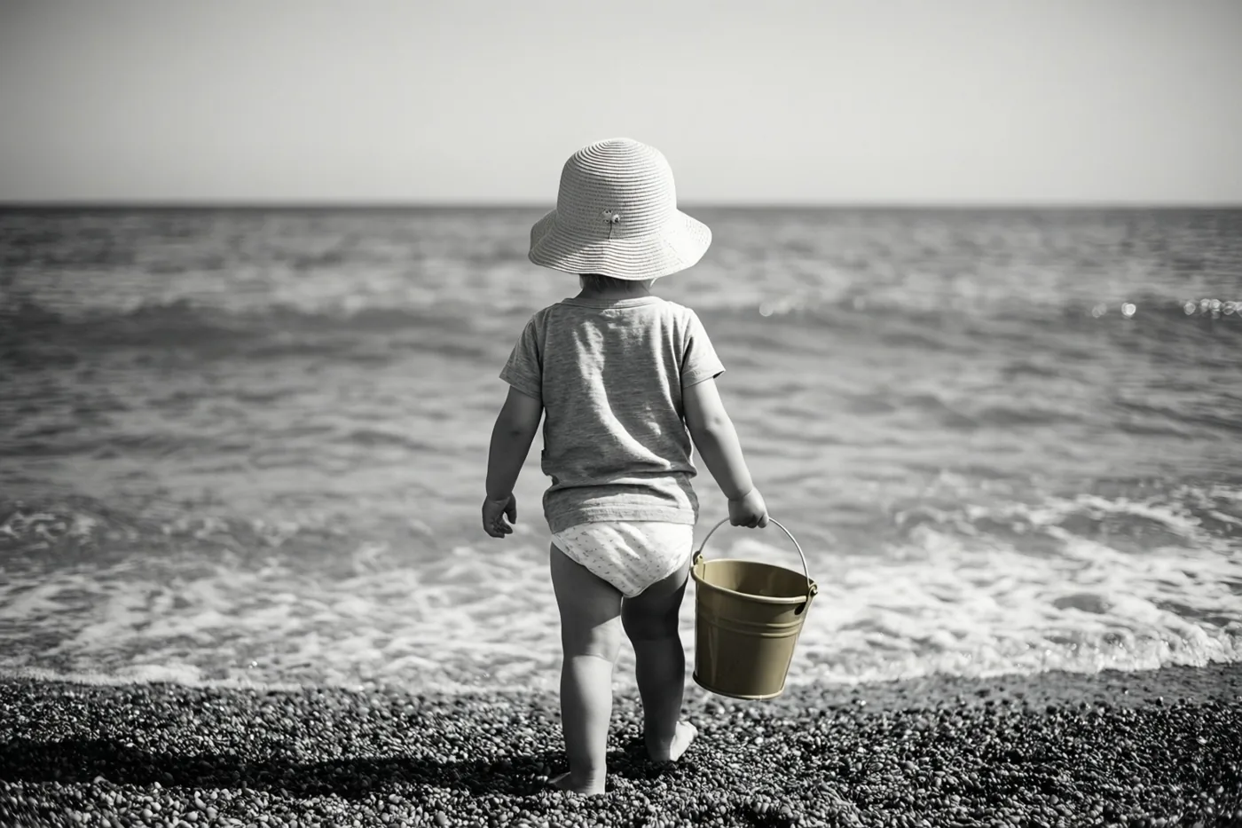 Een peuter met een zonnehoed loopt blootsvoets met een emmertje over een kiezelstrand naar de zee op een zwart-witfoto.