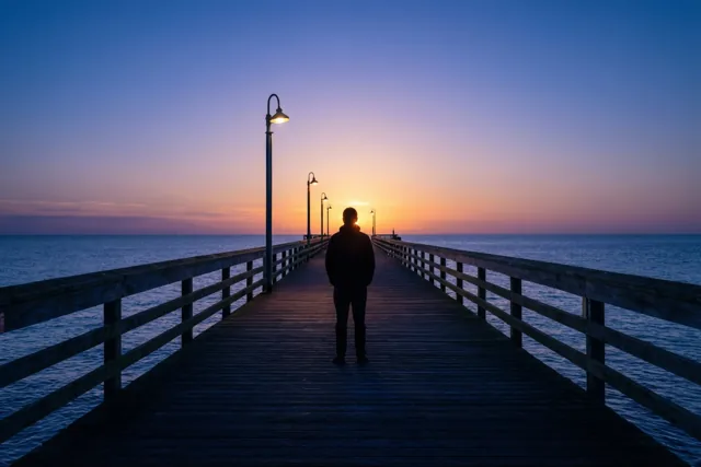 Een persoon staat aan het einde van een lange houten pier tegenover de opkomende zon boven zee, onder een blauwpaars-oranje hemel.
