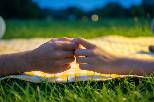 Twee handen met in elkaar gehaakte pinken rusten op een geel-wit geruit picknickdeken in groen gras, in warm licht voor een wazige blauwe avondlucht.