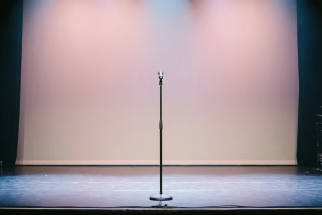 Een enkel microfoonstatief staat midden op een leeg concertpodium in zachtroze en blauw podiumlicht.