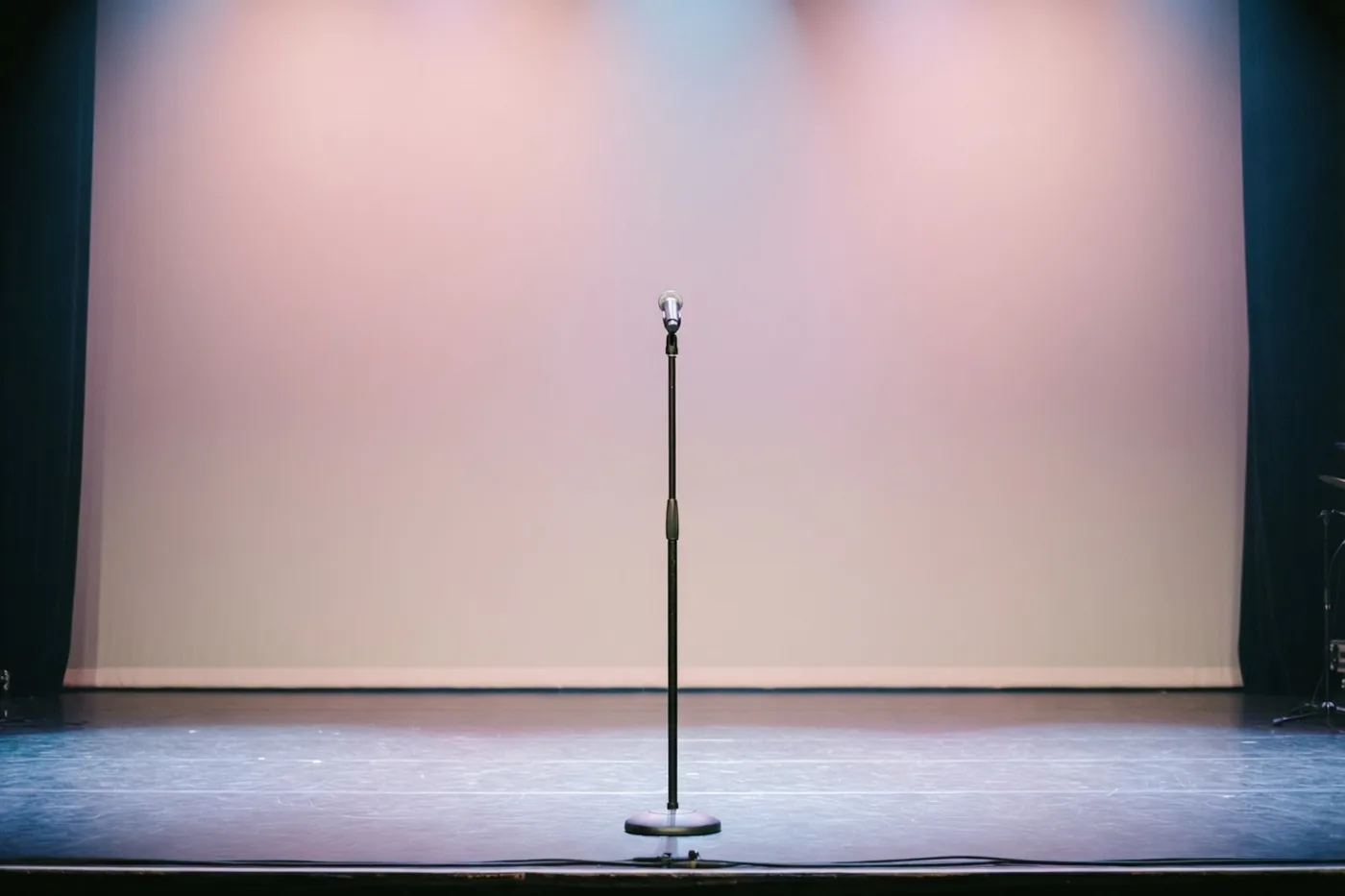 Een enkel microfoonstatief staat midden op een leeg concertpodium in zachtroze en blauw podiumlicht.