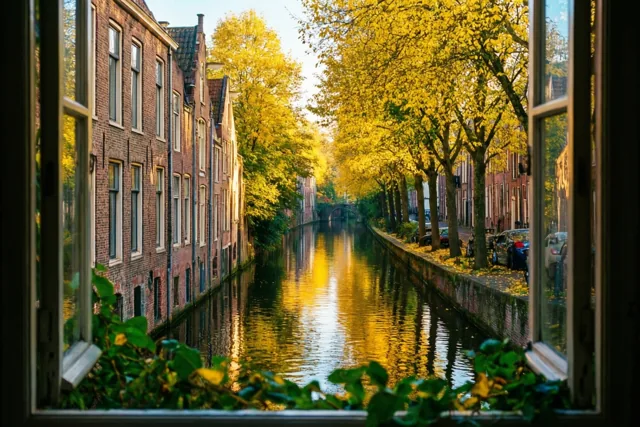 Door een open raam is een rustig kanaal te zien met oude bakstenen gebouwen en gouden herfstbomen die in warm zonlicht weerspiegelen in het water.