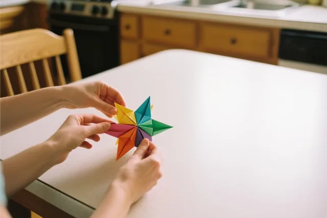 Handen vouwen een kleurrijke papieren sterlantaarn op een witte tafel in een keuken met houten kasten en een fornuis.