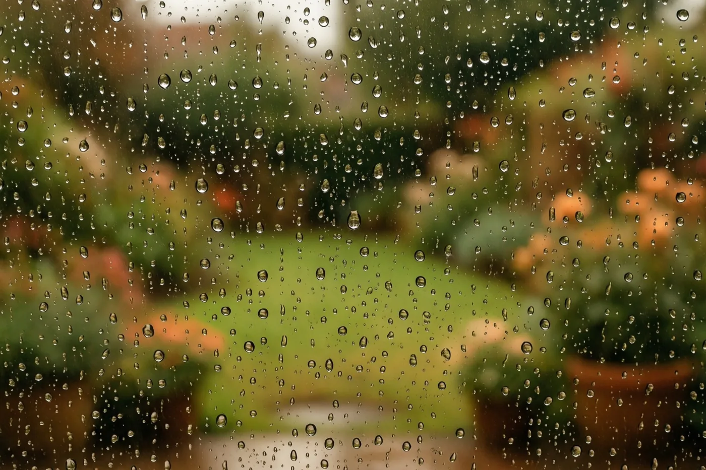 Een raam vol regendruppels vervaagt het zicht op een tuin in groene en bruine tinten.