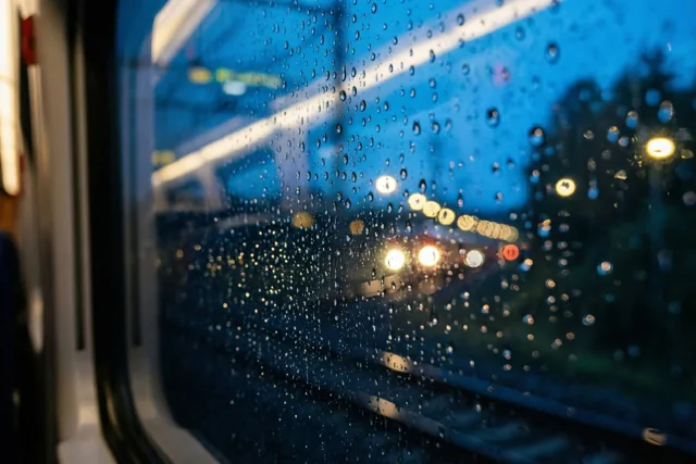 Regendruppels bedekken een treinraam en vervagen verre lichten tegen een donkerblauwe avondlucht.