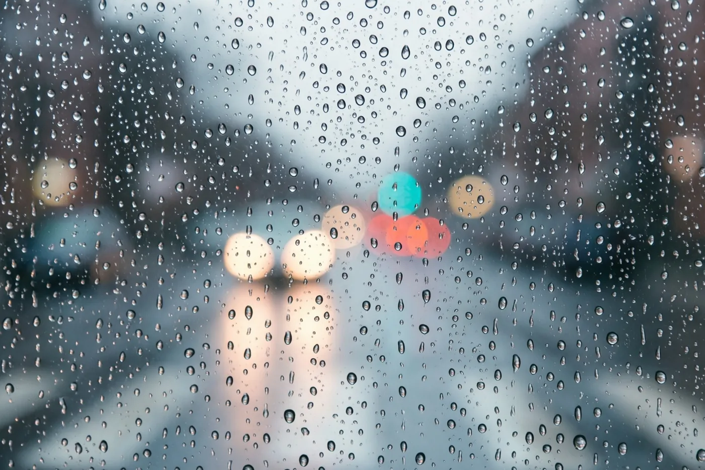 Een ruit is bedekt met regendruppels, met daarachter onscherpe straatlampen in koele tinten en zachte lichtvlekken.