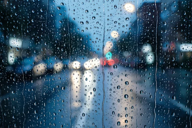 Regendruppels lopen over een ruit en vervagen de straatlampen en stadslichten erachter, in koele blauwe en grijze tinten.