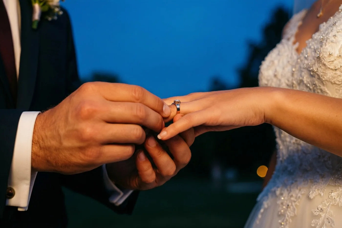 Een close-up toont hoe een bruidegom een ring aan de vinger van de bruid schuift, met warm licht op de handen en haar witte kralenjurk tegen een diepblauwe schemerhemel.