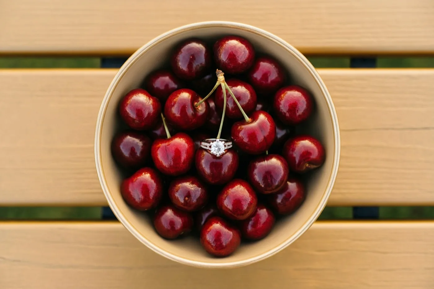 Een kom glanzende rode kersen staat op een houten picknicktafel met een verlovingsring die in het midden aan een kersensteeltje hangt.