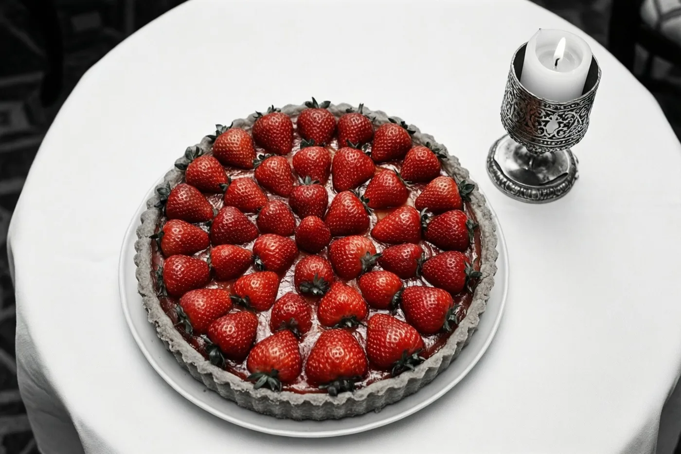Een taart met hele rode aardbeien staat op een wit tafelkleed naast een sierlijke zilveren houder met een brandende witte kaars, waarbij de rest van het beeld zwart-wit is.