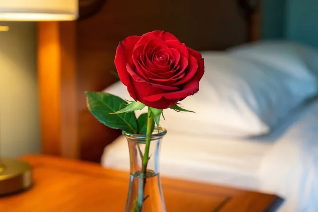 Een rode roos in een smalle glazen vaas staat op een houten nachtkastje, met een zacht verlicht bed en een lamp op de achtergrond.