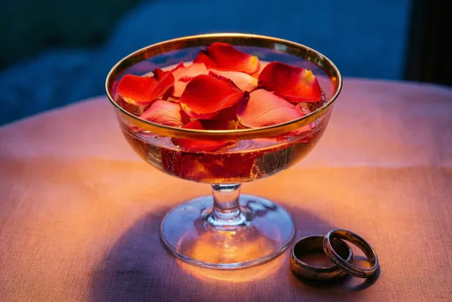 Een champagnecoupe met gouden rand en rode rozenblaadjes staat naast twee in elkaar gehaakte trouwringen op een linnen tafelkleed tegen een donkerblauwe achtergrond.