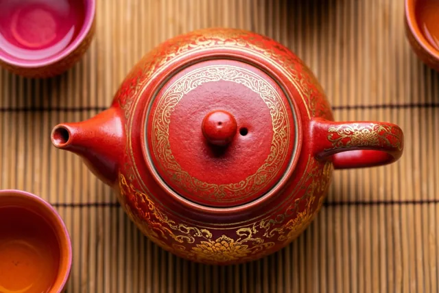 Een rode porseleinen theepot met gouden patronen staat tussen twee bijpassende kopjes op een bamboemat.
