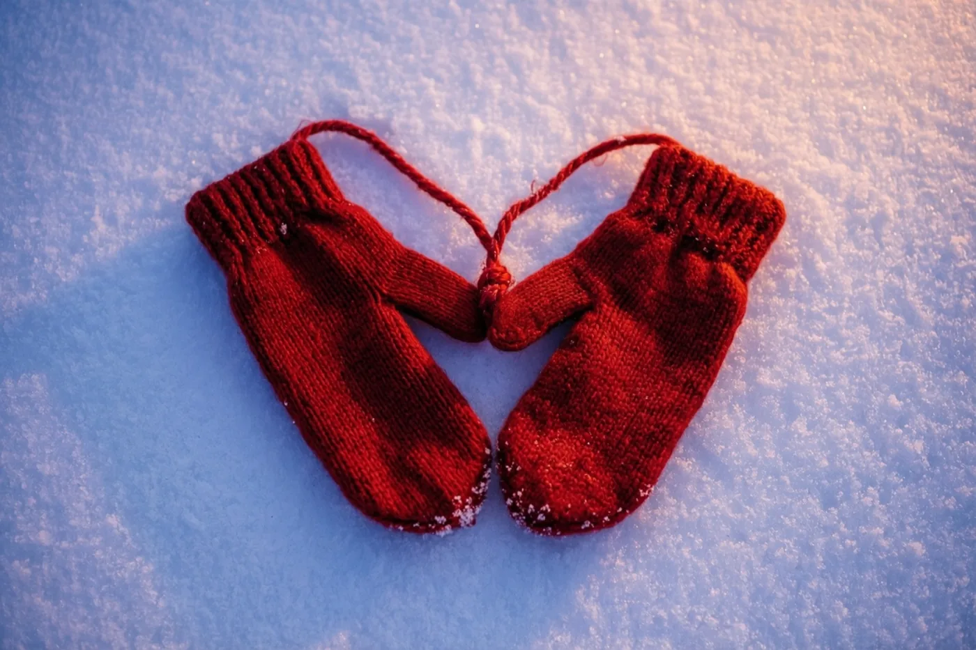 Twee kleine rode gebreide wantjes liggen in de vorm van een hart op verse sneeuw en zijn verbonden door een rood koordje, tegen een blauw-witte achtergrond.