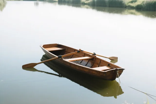 Een houten roeiboot met twee roeispanen ligt aangemeerd op een kalme vijver, met riet langs de verre oever in zacht licht.