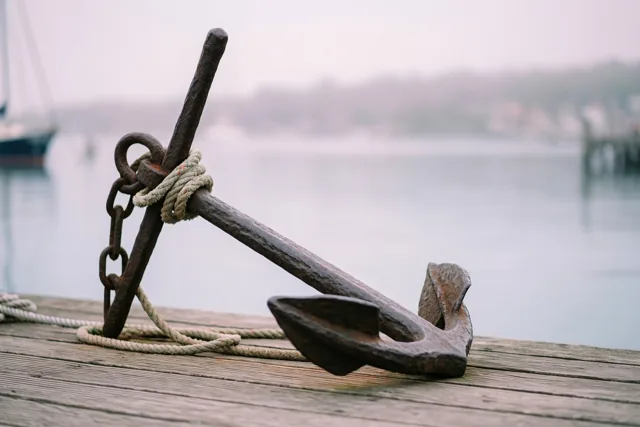 Een roestig ijzeren anker met dik touw rond de schacht ligt op een verweerde houten aanlegsteiger, met mistig water, een wazige zeilboot en een verre oever erachter.