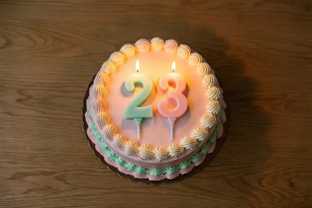 Een ronde roomtaart met opgespoten versiering en brandende cijferkaarsjes die 23 vormen staat op een houten tafel met zachte schaduwen.