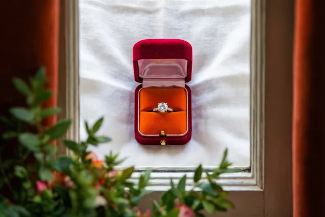 Een open rood fluwelen doosje met een diamanten verlovingsring staat midden op een witte linnen doek, omkaderd door een raamachtige structuur met wazig groen op de voorgrond.