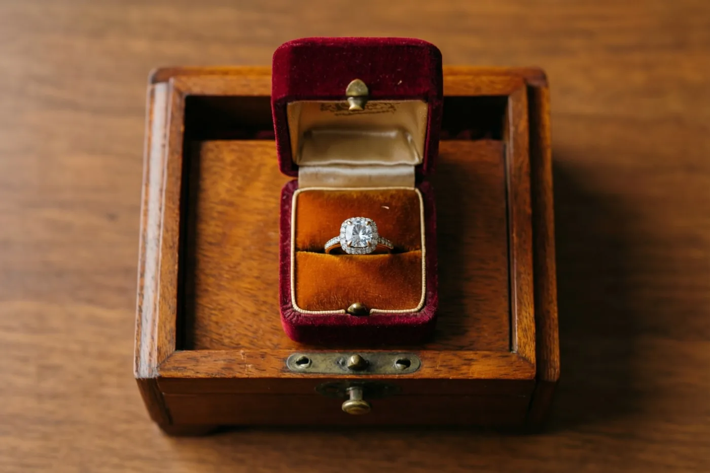 Een ring met één diamant ligt in een rood fluwelen ringdoosje op een vintage houten muziekdoos, op een houten ondergrond in warme tinten.