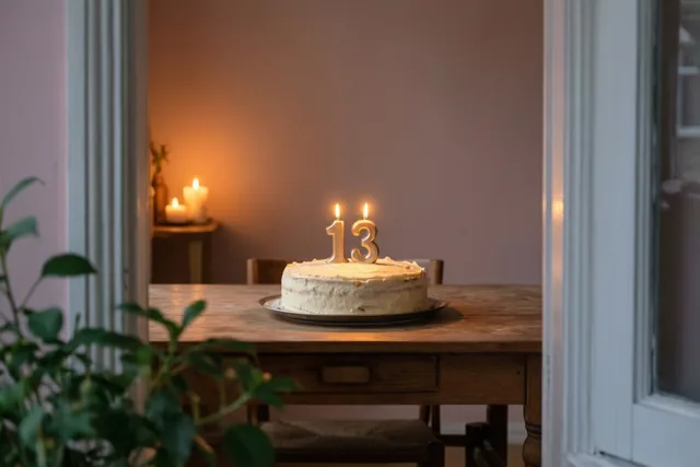 Door een deuropening is een zelfgemaakte roomtaart te zien met brandende cijferkaarsjes die 13 vormen, op een houten tafel in warm kaarslicht.