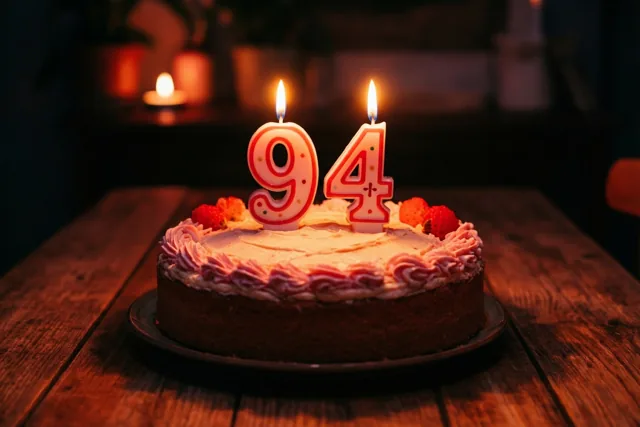 Een zelfgemaakte roomtaart met brandende cijferkaarsjes 94 staat op een houten tafel in warme oranje, bruine en roze tinten.