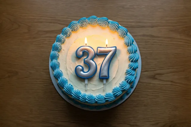 Een ronde roomtaart met blauw opgespoten glazuur en brandende cijferkaarsjes 37 staat op een houten tafel.