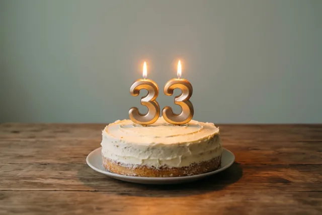 Een zelfgemaakte roomtaart met twee brandende gouden cijferkaarsen die 33 vormen staat op een wit bord op een houten tafel.