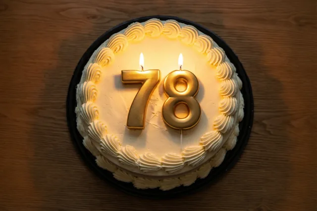 Een ronde roomtaart met gespoten versiering en twee brandende gouden cijferkaarsen die 78 vormen, staat op een houten tafel.