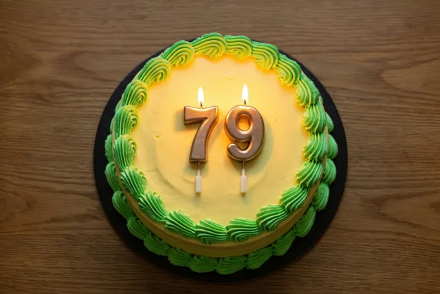 Een ronde roomtaart met groene gespoten versiering en brandende gouden cijferkaarsen 79 staat op een houten tafel.