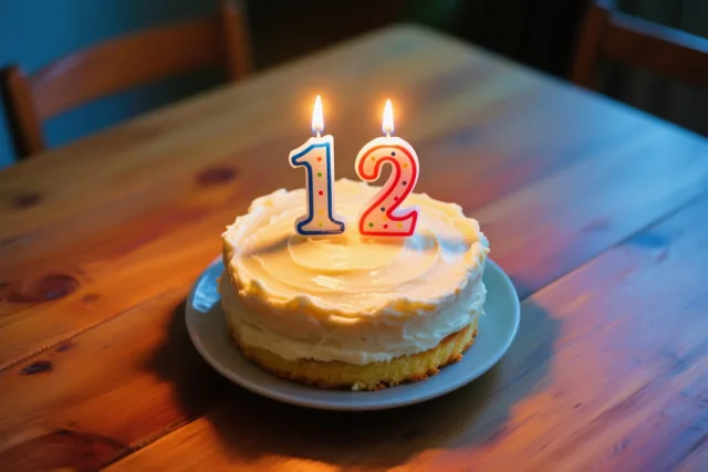 Een zelfgemaakte roomtaart met twee brandende cijferkaarsen die samen 12 vormen staat op een houten tafel in warm kaarslicht.
