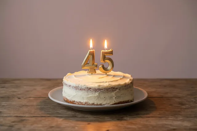 Een huisgemaakte roomtaart met brandende cijferkaarsjes 45 staat op een wit bord op een houten tafel in warm kaarslicht.