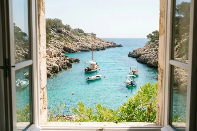 Door een raam met een stenen omlijsting is een rotsachtige baai met turquoise water en kleine aangemeerde boten te zien, met groen op de voorgrond.