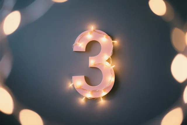 Een roze cijfer 3 is omrand met warme lichtjes tegen een donkere muur, omgeven door wazige feestlichtjes.