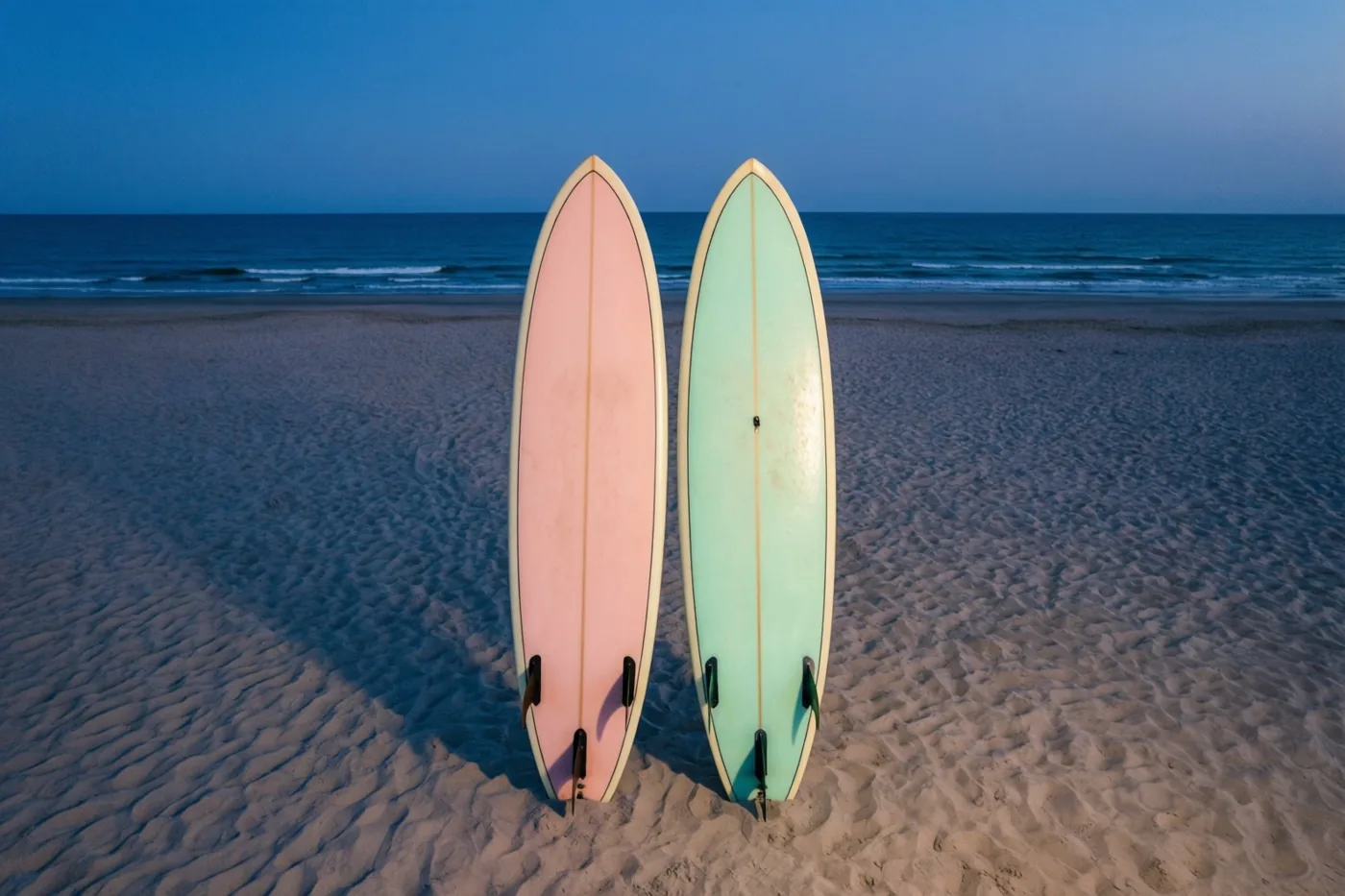 Een roze en een lichtgroene surfplank staan rechtop in het zand op een breed strand, met de zee erachter in het zachte blauwe licht van de schemering.