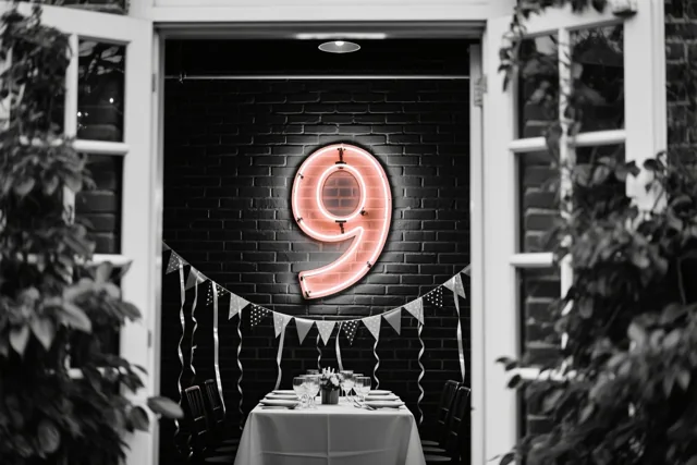 Een grote roze neonnegen hangt aan een zwarte bakstenen muur boven een gedekte feesttafel met zwart-witte slingers, gezien door open witte deuren met groen eromheen.