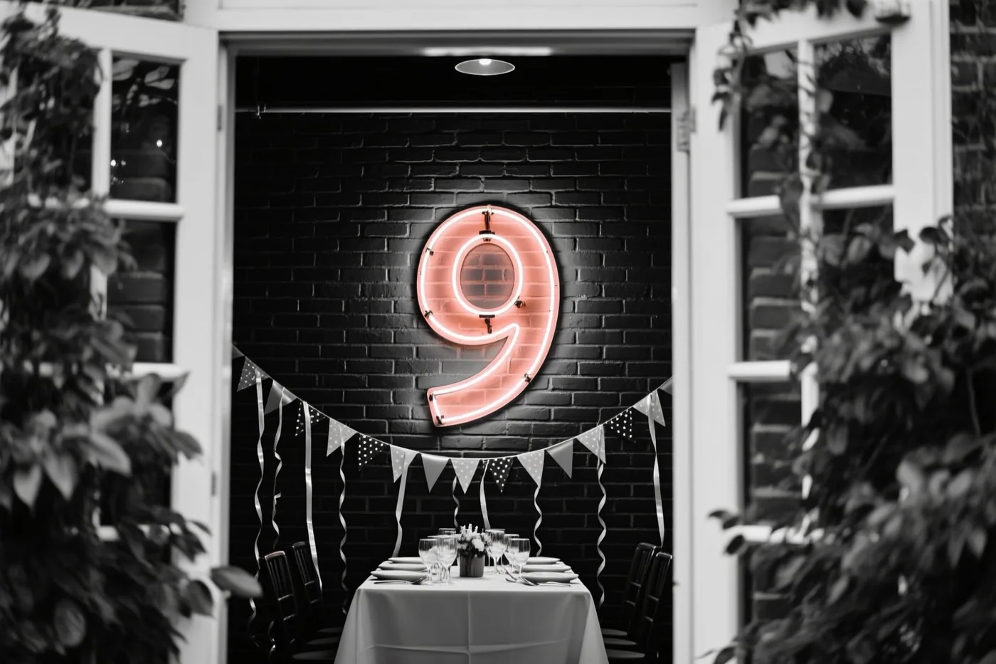 Een grote roze neonnegen hangt aan een zwarte bakstenen muur boven een gedekte feesttafel met zwart-witte slingers, gezien door open witte deuren met groen eromheen.