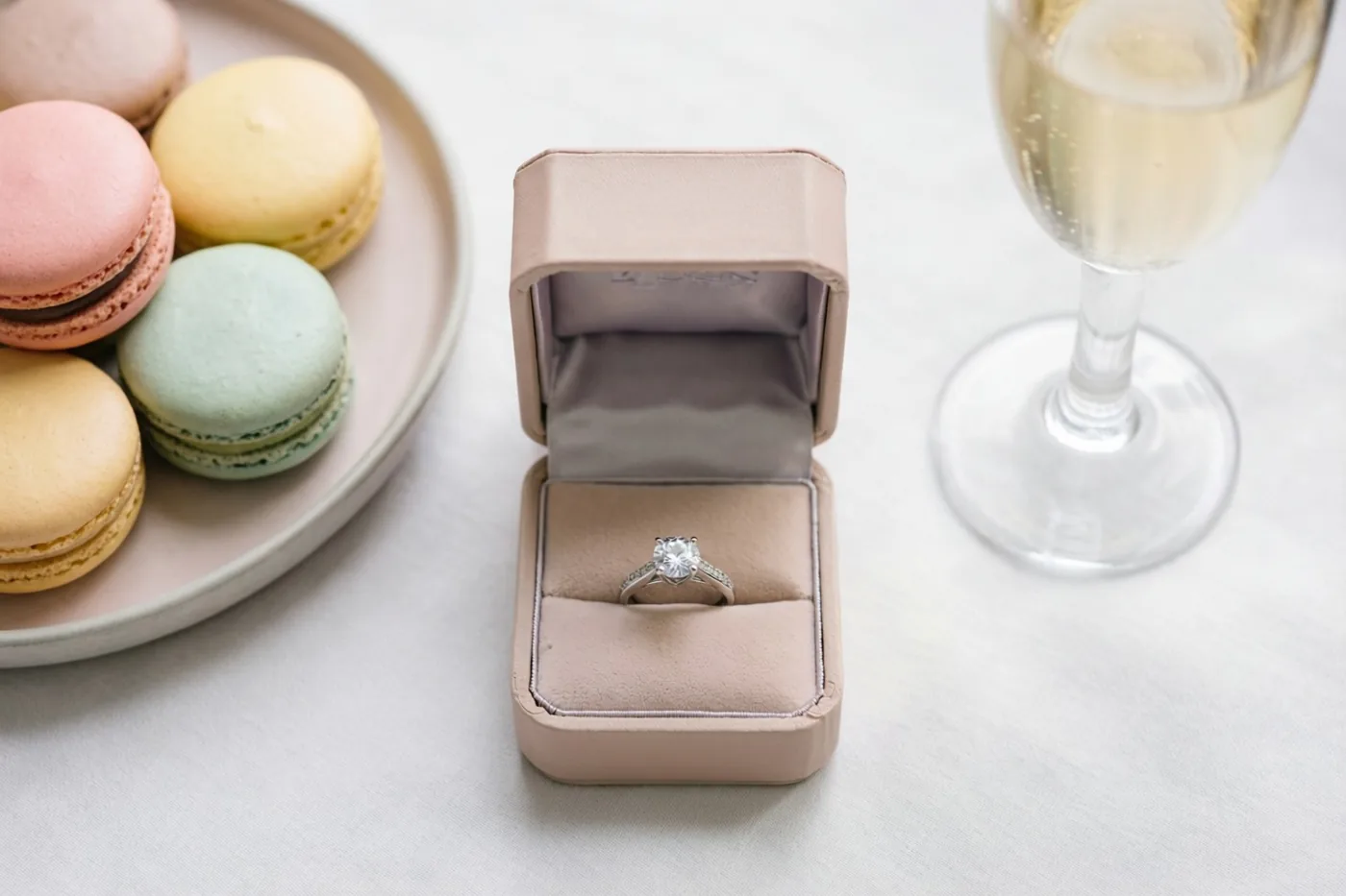 Een open lichtroze ringdoosje met een diamanten ring staat op een wit tafellaken naast een bord macarons in pasteltinten en een glas champagne.
