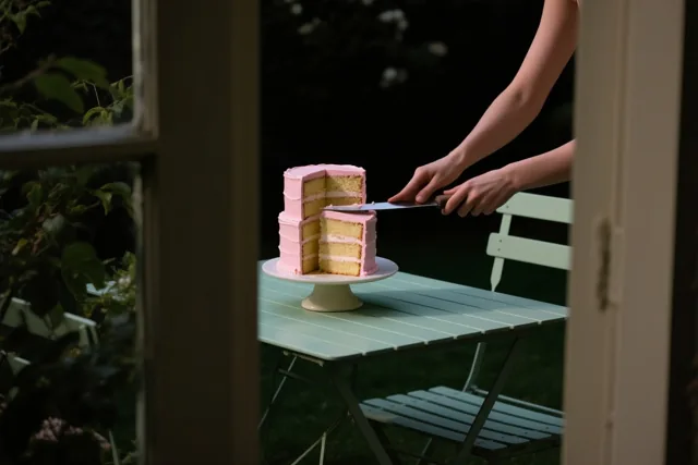 Door een raamkozijn zijn handen te zien die het eerste stuk uit een hoge laagjestaart met roze glazuur snijden op een lichtgroene tuintafel, met groen op de achtergrond.