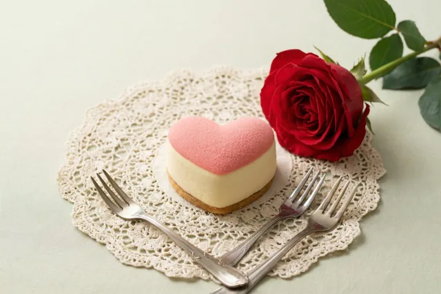 Een hartvormig taartje met een roze bovenkant en een romige onderkant staat op een wit kanten onderleggertje, met een rode roos en twee dessertvorkjes op een lichtgroen tafelkleed.