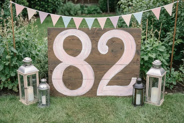 Een houten bord met 82 in lichtroze verf staat op het gras tussen lantaarns, onder een slinger met pastelkleurige vlaggetjes en met groen erachter.