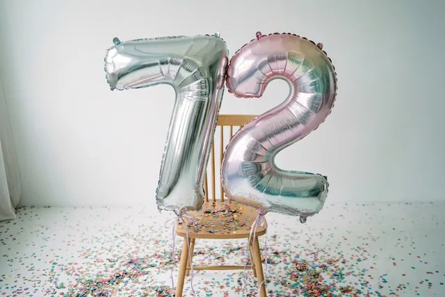 Zilveren en pastelroze folieballonnen in de vorm van 7 en 2 hangen aan een houten stoel in een lichte kamer met kleurrijke confetti op de vloer.
