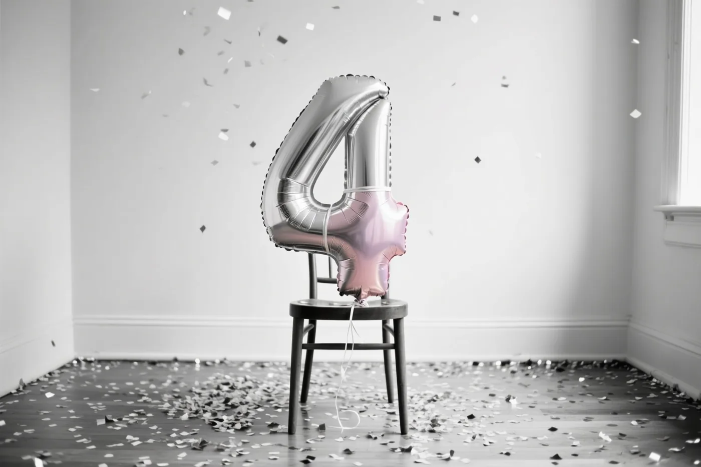 Een zilveren en pastelroze cijferballon 4 hangt aan een houten stoel in een lichte kamer, met zilveren confetti in de lucht en op de vloer.