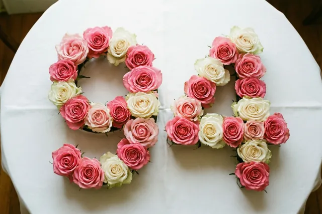 Van bovenaf gezien vormen roze en witte rozen het getal 94 op een wit tafelkleed in zachte pasteltinten.