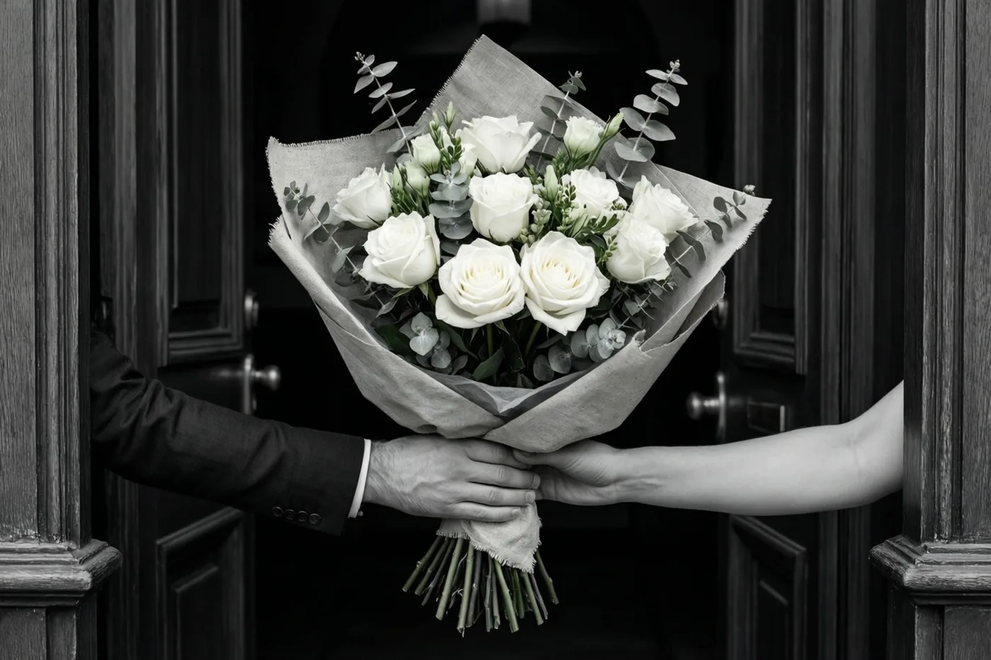 Twee handen, één uit een kostuummouw en één met een blote arm, geven een boeket witte rozen en eucalyptus in linnenpapier door een open houten deur, op een verder zwart-witte foto.