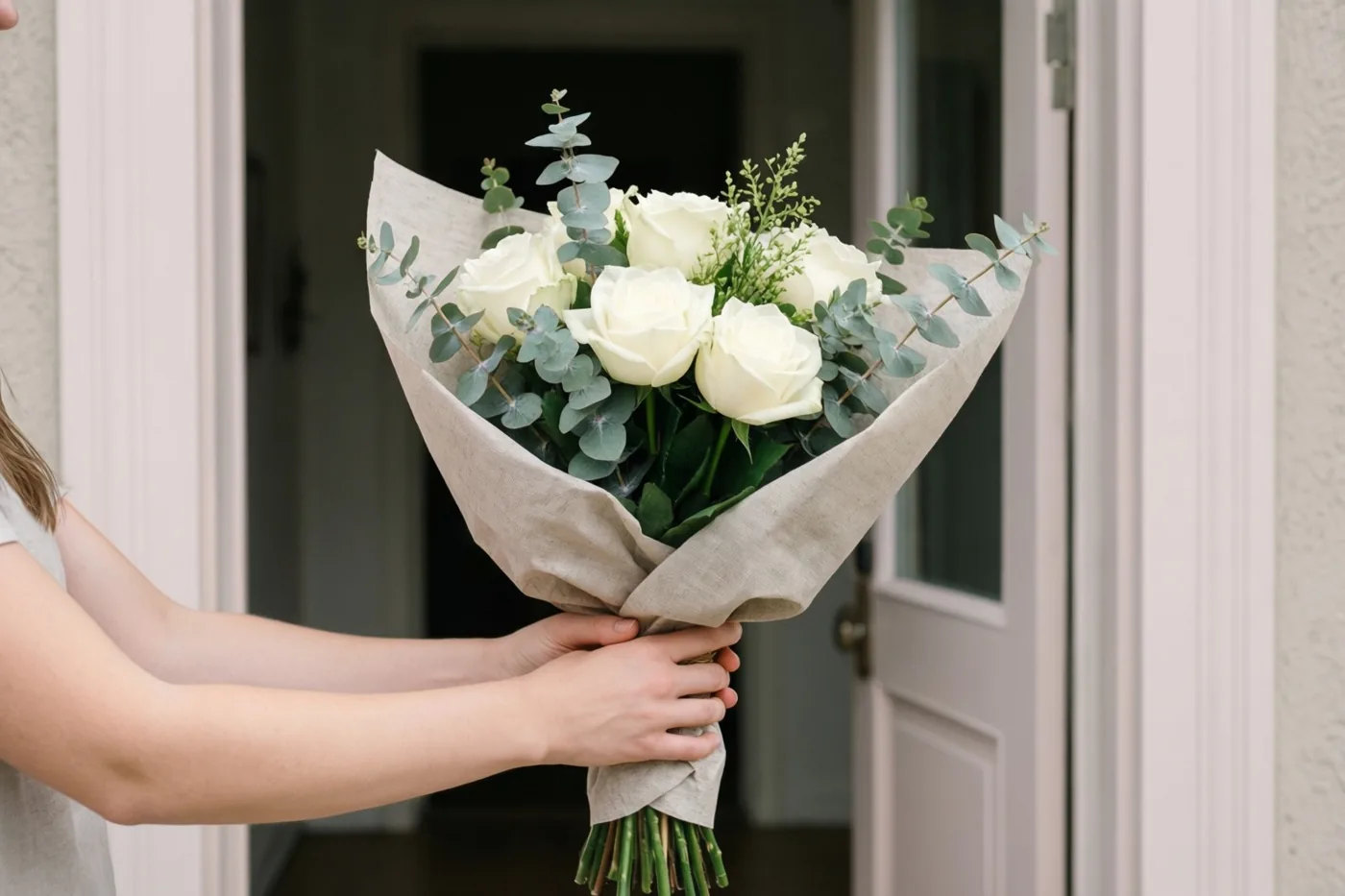 Handen houden aan een open voordeur een boeket witte rozen en eucalyptus in beige linnenpapier vast in zacht licht.