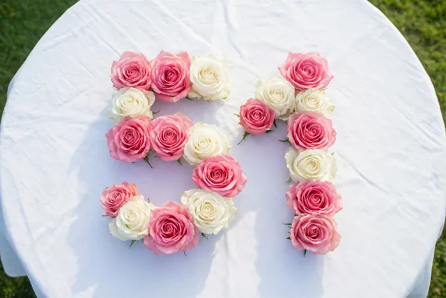Roze en witte rozen vormen het getal 51 op een wit tafelkleed buiten op het gras, van bovenaf gefotografeerd in zacht daglicht.