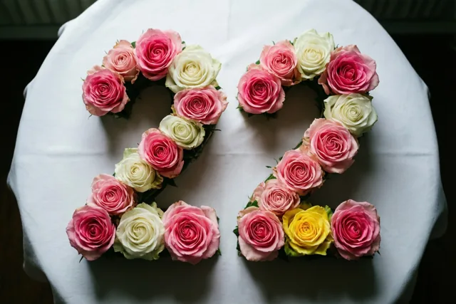 Van bovenaf zijn roze en witte rozen te zien die 22 vormen op een wit tafelkleed, met zacht licht op de bloemblaadjes.