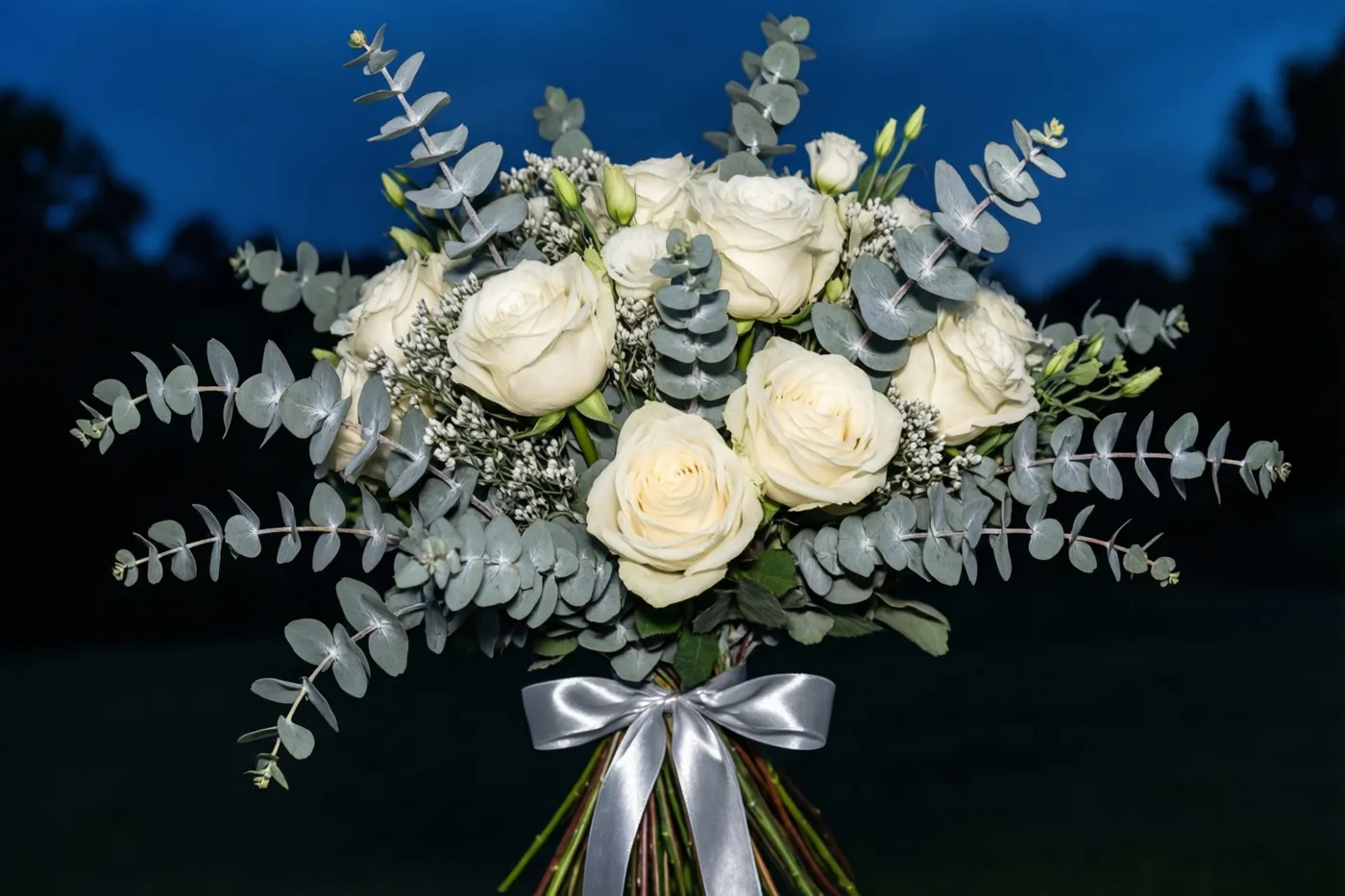 Een boeket witte rozen en zilvergrijze eucalyptus met een zilveren lint staat afgebeeld tegen een donkerblauwe avondhemel.
