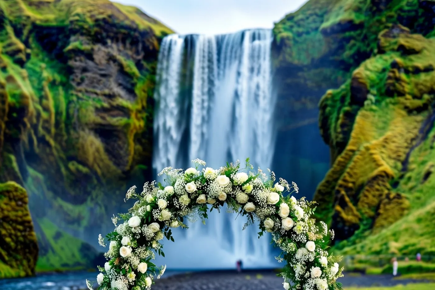 Een boog met witte rozen en gipskruid staat in een groene vallei voor een grote waterval die over met mos begroeide rotswanden stroomt.